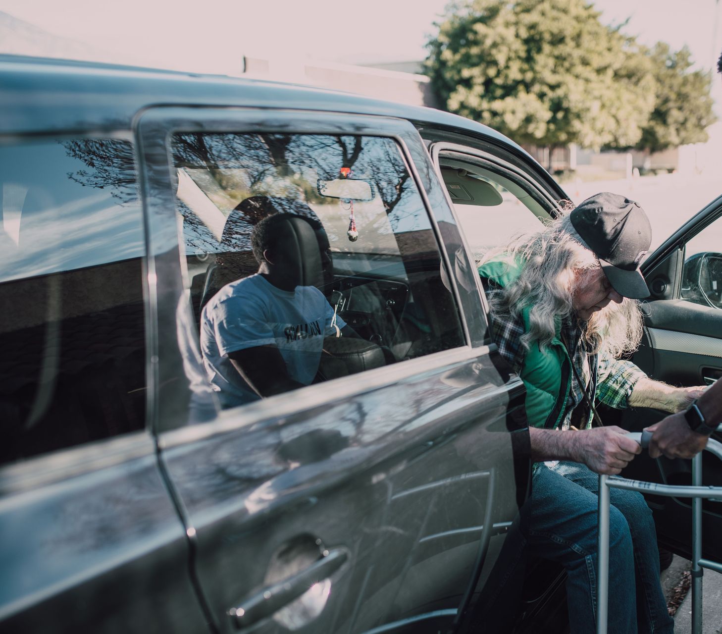 Two men help an elderly person with a walker get out of a car outdoors.